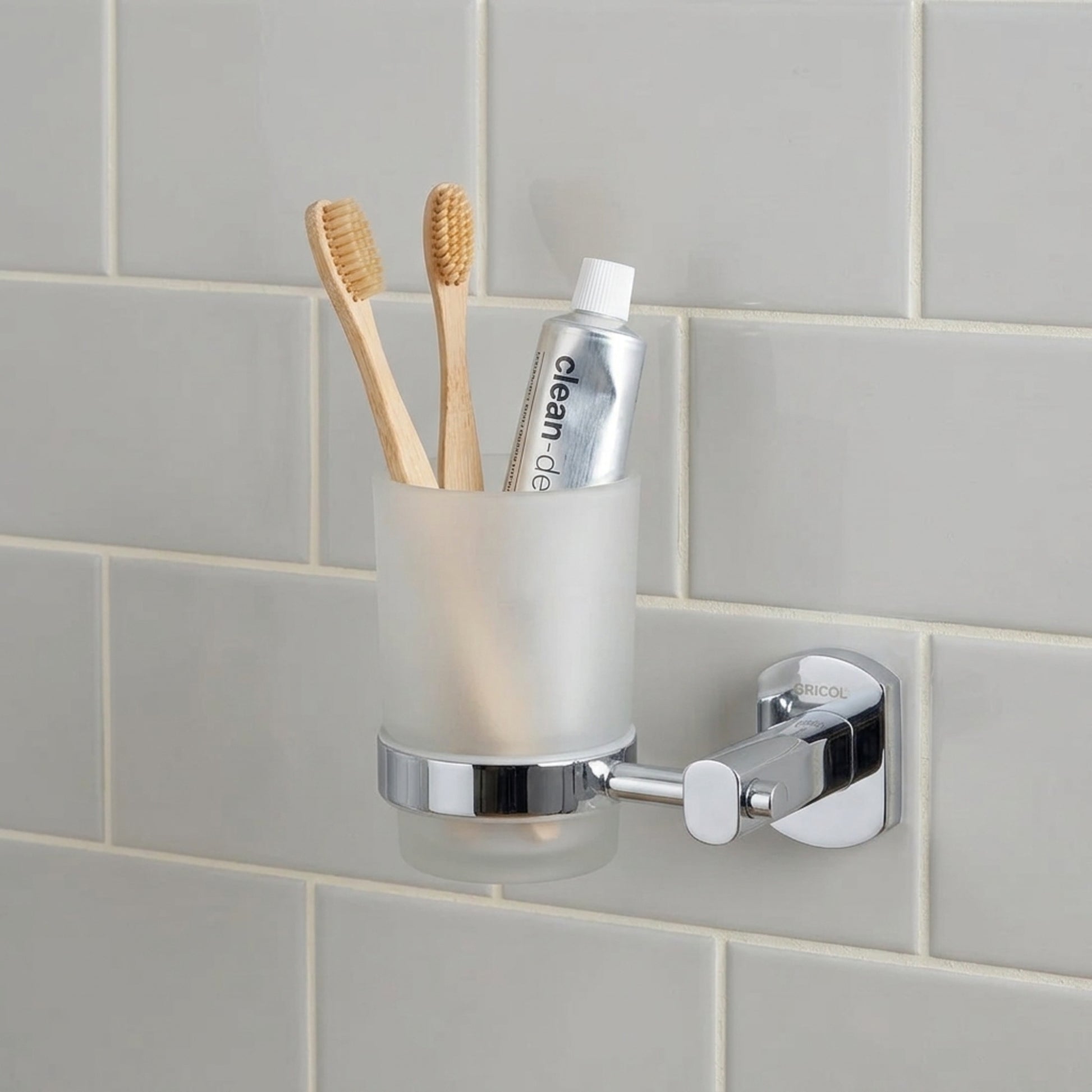Portacepillos vaso en vidrio y soporte metálico Portobello para baño GRICOL en acabado cromado instalado en la pared de un baño con cepillos y pasta dental.